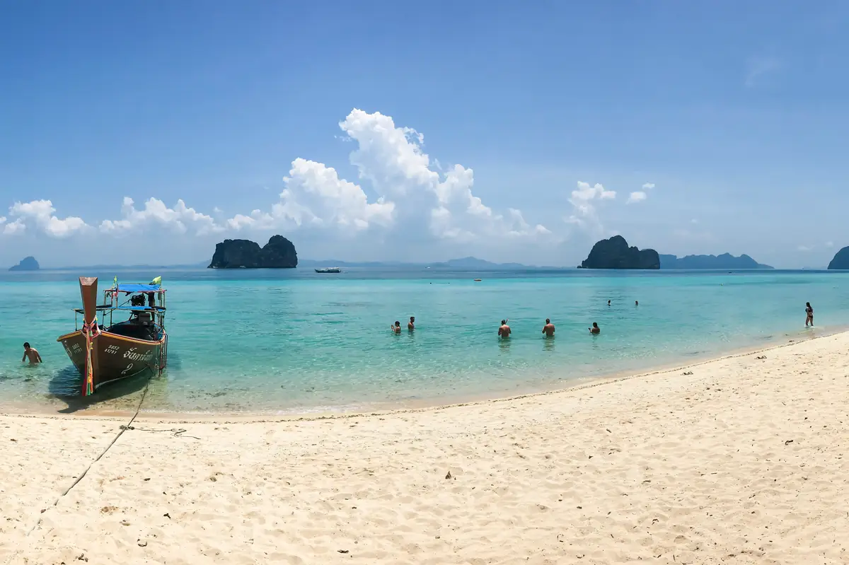 a white sandy beach, clear sunny sky, clear calm water, people are entering to go snorkeling, small islands on the horizon. A traditional wooden fishing boat is moored on the sand on the right of the picture.