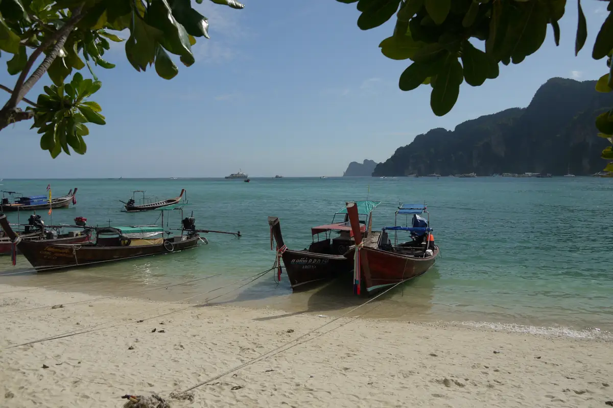wooden longtail boats are moored on the sand of Tonsai bay, Phi Phi Island. Ccliffs rise on the other side of the bay. On the horizon are speedboats and larger ferry boat.